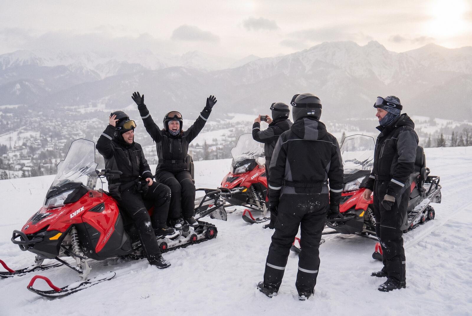 A couple in helmets with arms raised on a snowmobile at a viewpoint, the rest of the group beside them, Zakopane in the valley below and the Tatras on the horizon