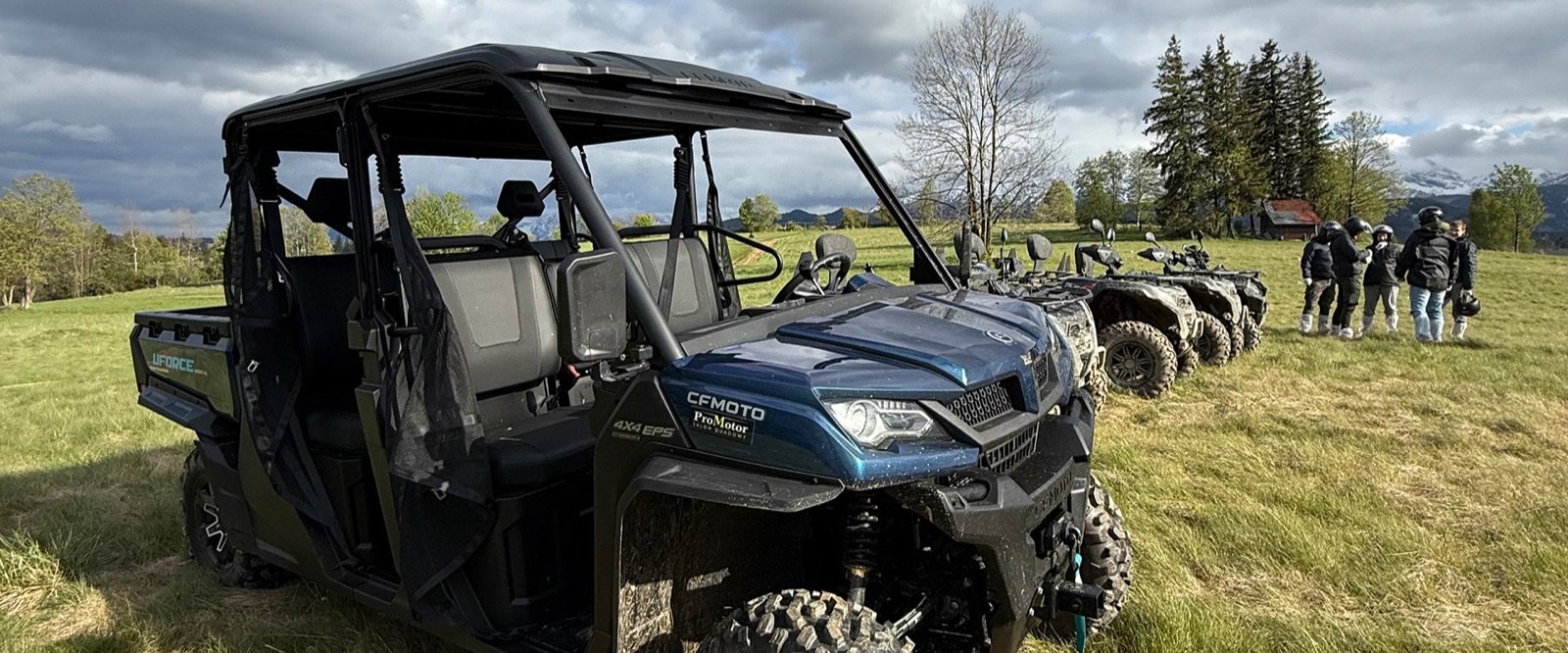 A six-seater buggy on a meadow with quads and a group beside it, clouds over the mountains behind