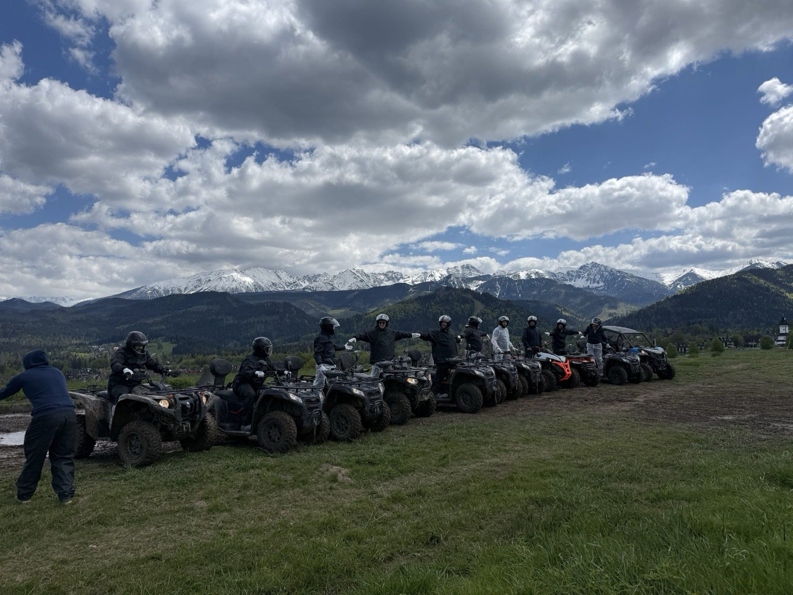 A row of quads with riders on a meadow, snow-capped Tatra peaks behind