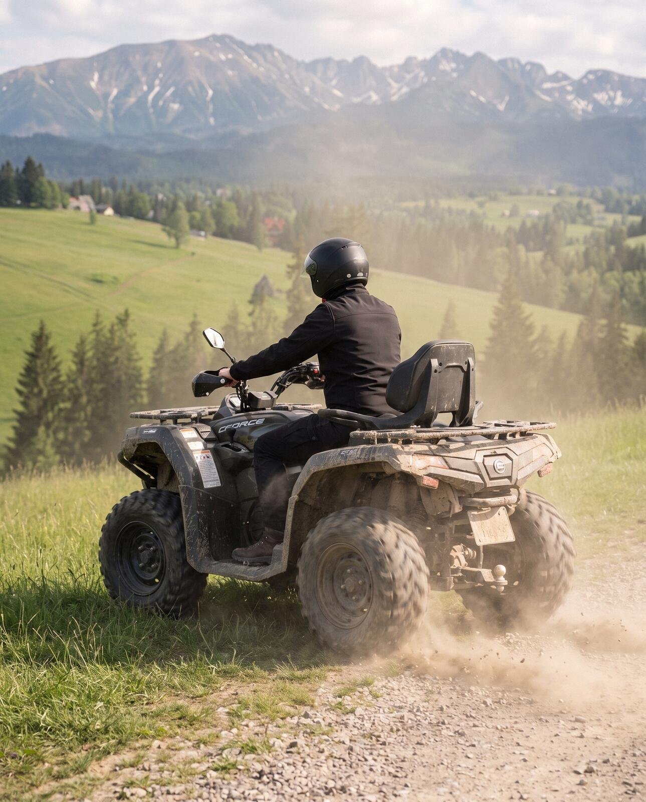 A quad seen from behind kicking up dust on a grassy bend, meadows, spruces and the Tatras ahead