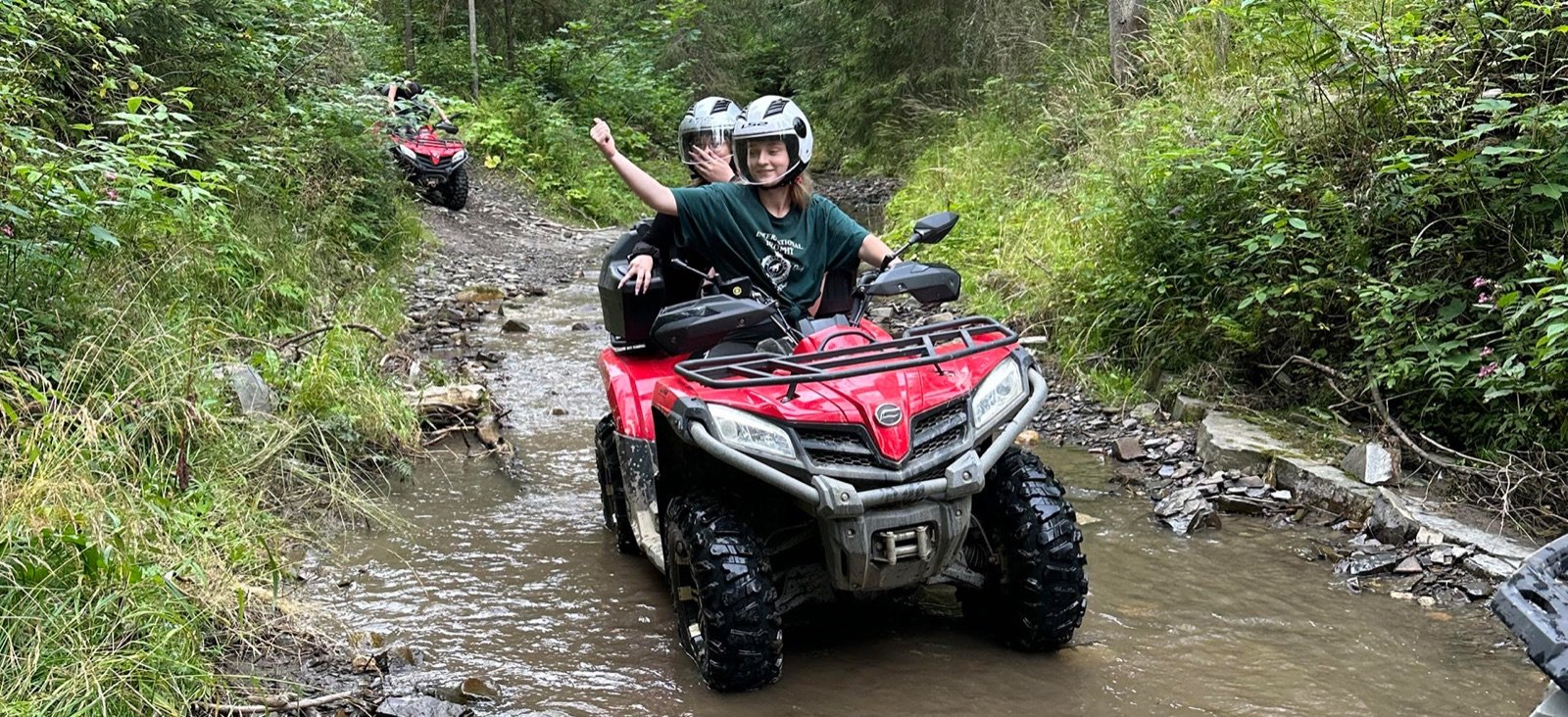 Two people on a red quad crossing a mountain stream in the forest