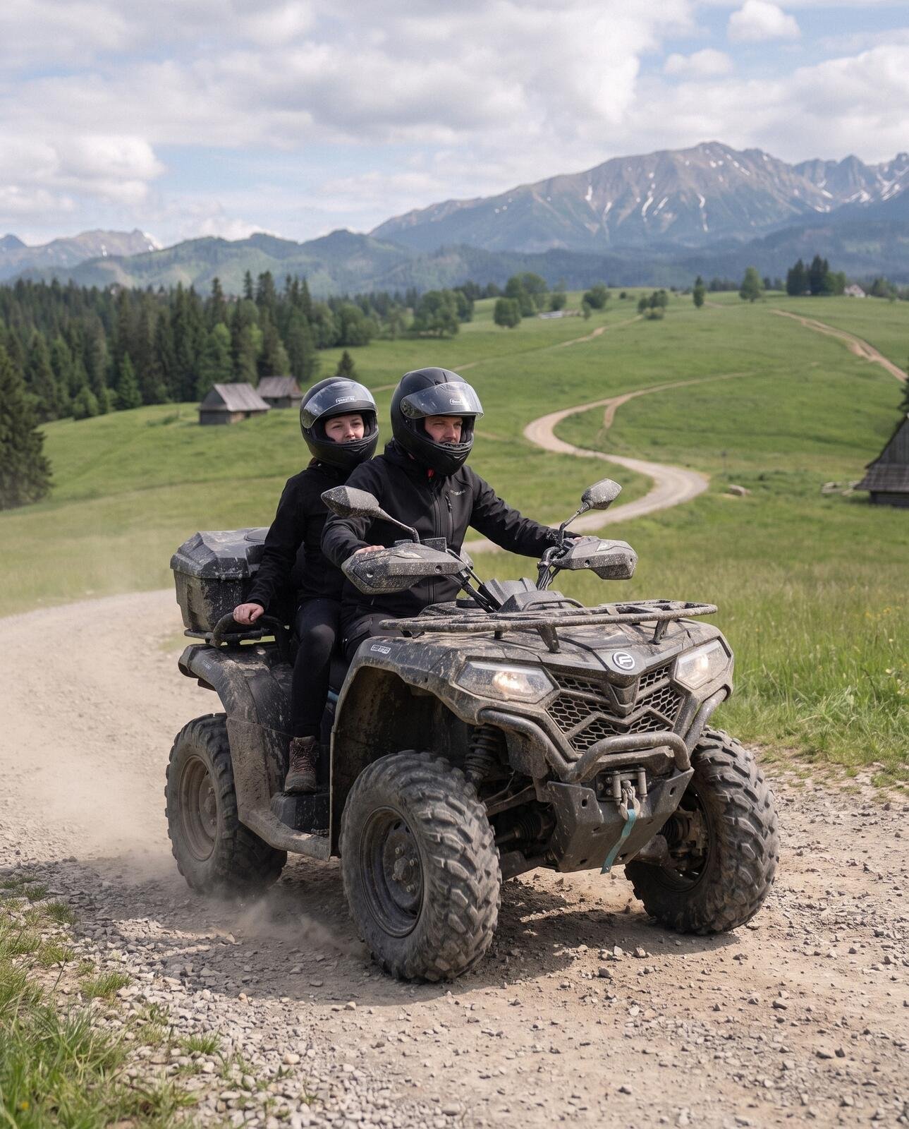 Two people in helmets on one quad on a gravel track, a winding road through meadows with wooden huts and the Tatras behind