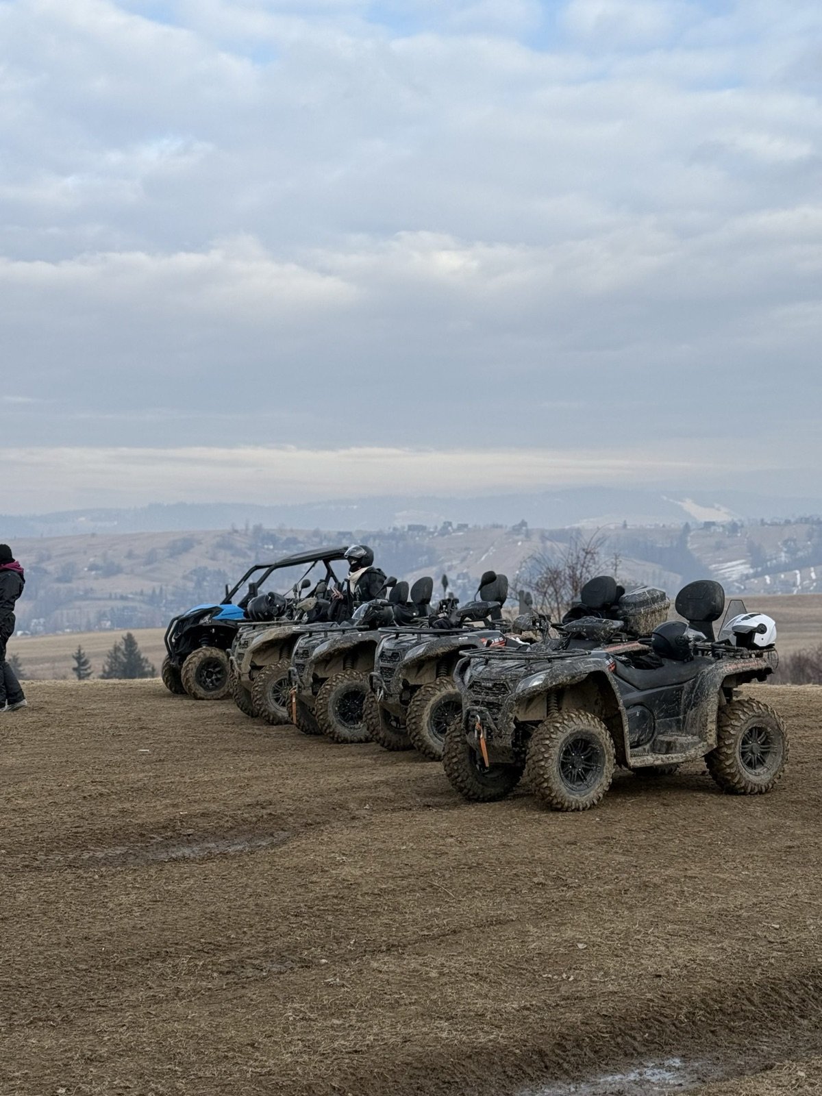 Quads and a buggy lined up on a ploughed field, a mountain range behind under an overcast sky