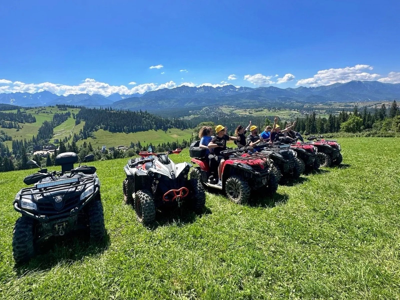 A row of quads with a group in yellow caps on a green meadow, the full Tatra panorama behind on a sunny day