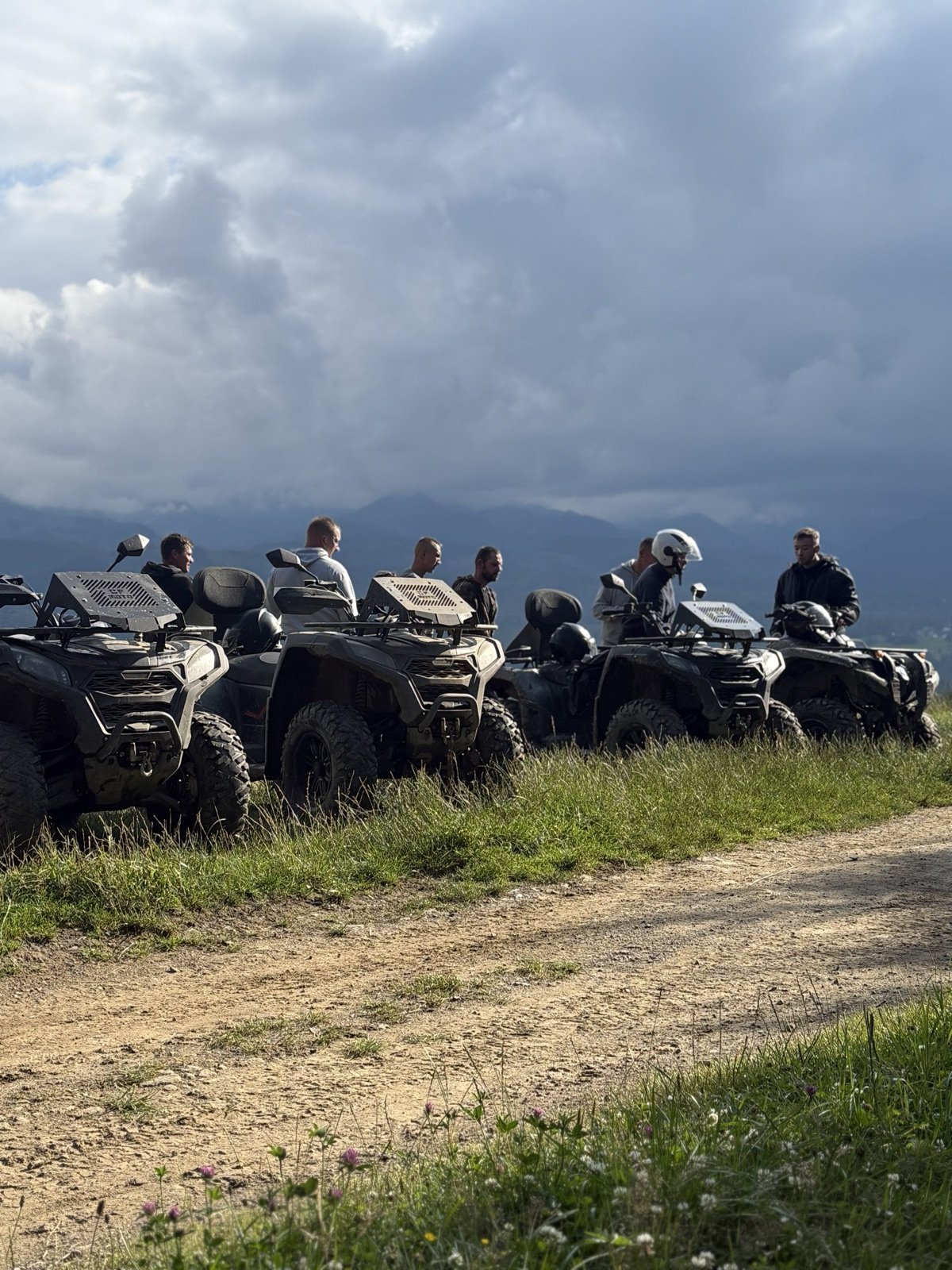 A group on quads heading down a dirt track under a dark, stormy sky