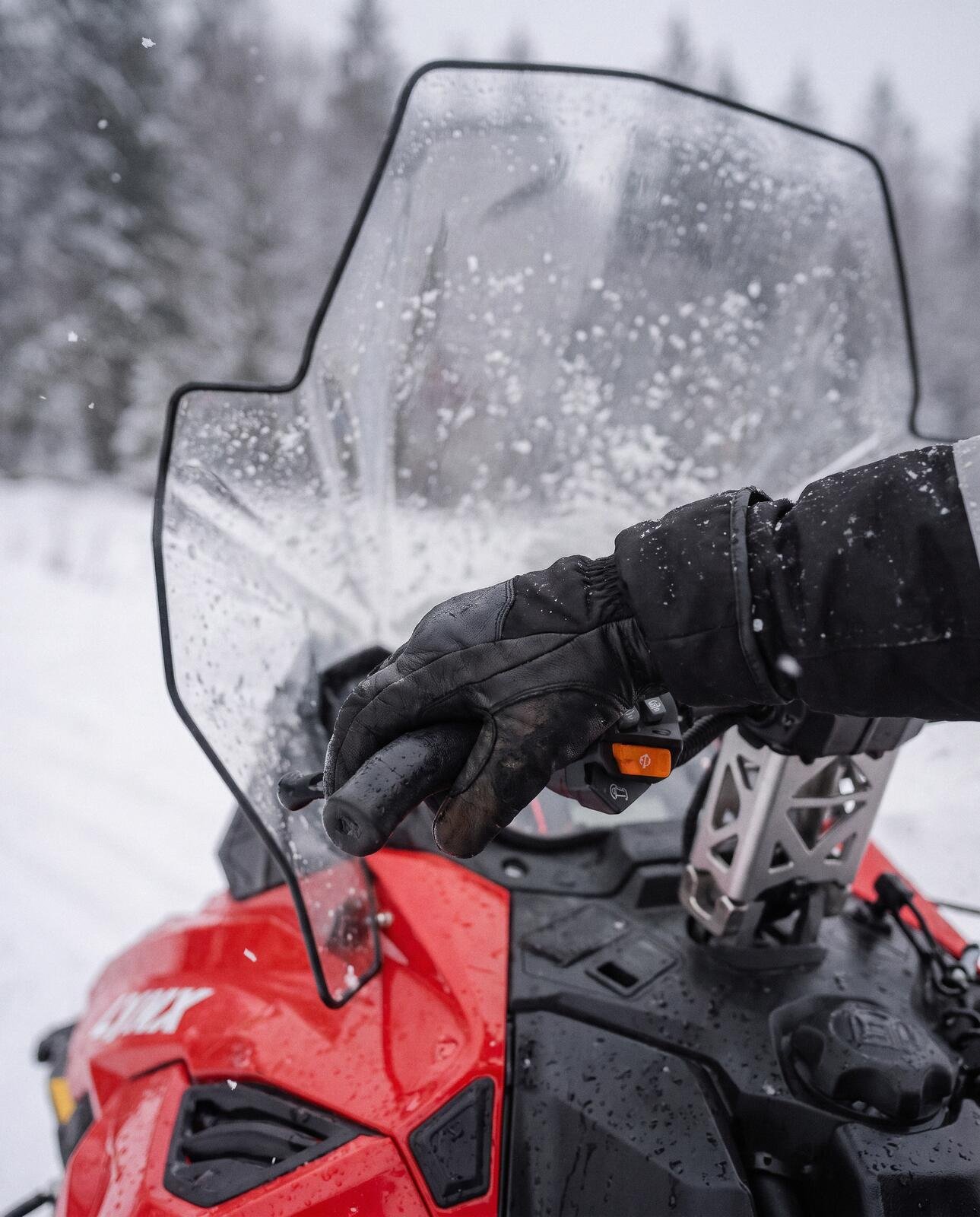 A gloved hand on the throttle of a red snowmobile, snowflakes on the windscreen and a winter forest behind