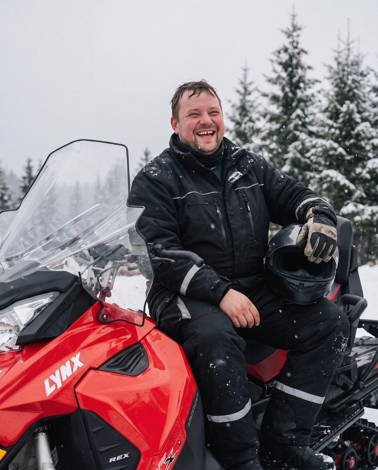 A laughing rider in a winter suit sitting on a red snowmobile with a helmet in hand, snow-covered spruces behind