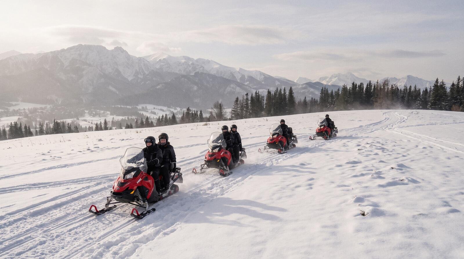 A group in helmets riding snowmobiles in a line up a snowy slope above Zakopane, the snow-capped Tatra panorama behind
