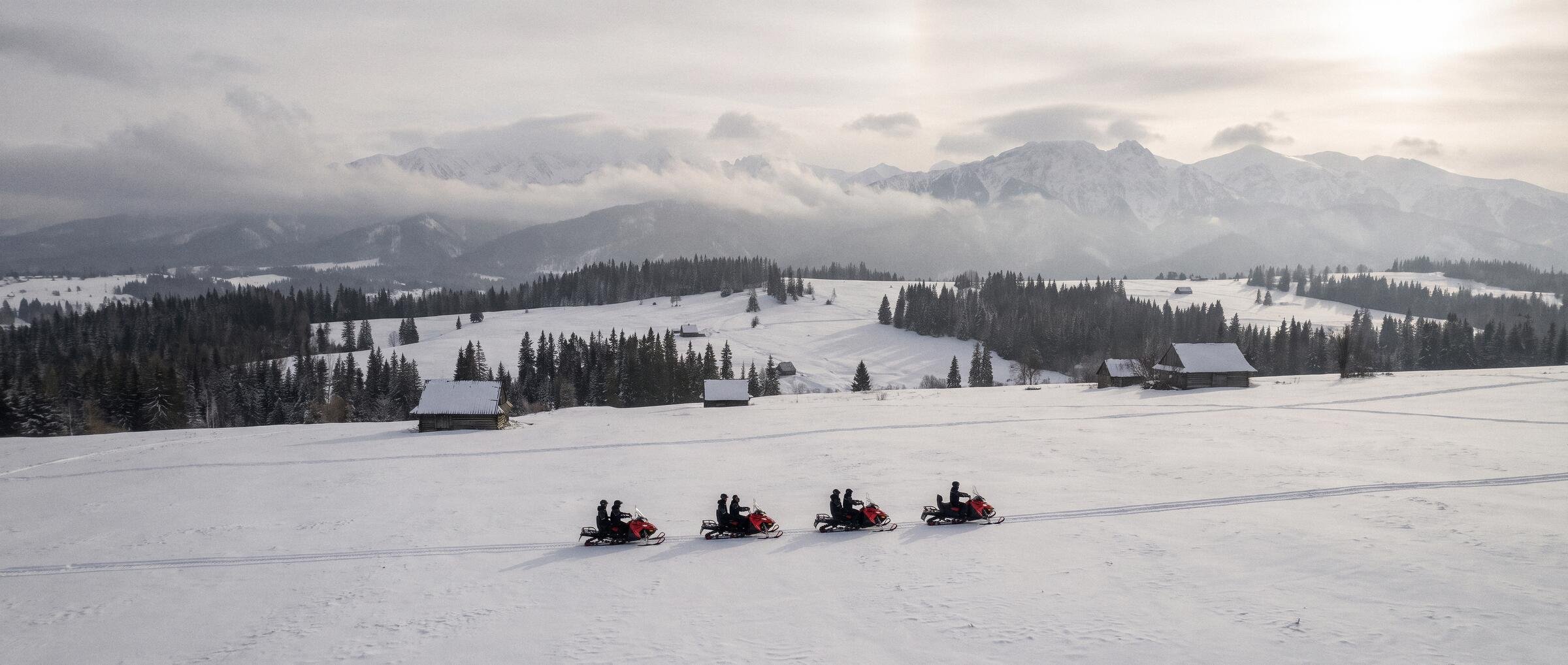 Four snowmobiles crossing a snowy clearing above Zakopane, wooden huts, spruce forest and the full Tatra panorama behind