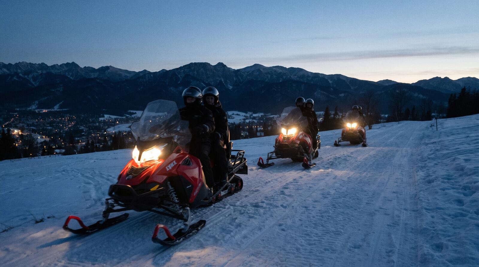 Snowmobiles with their headlights on riding a snowy track at dusk, the lights of Zakopane below and the Tatras on the horizon