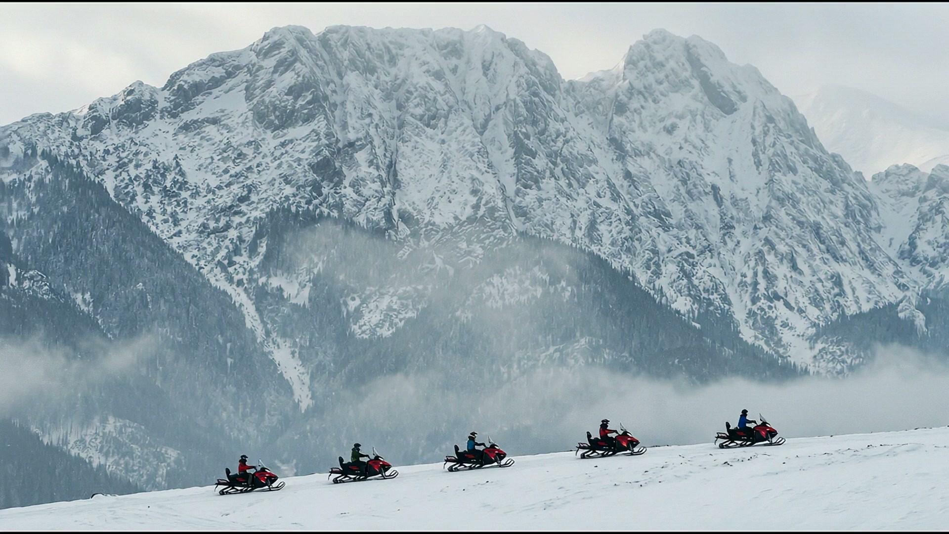 Five snowmobiles lined up on a snowy meadow, the snow-covered face of Giewont rising in the mist behind them